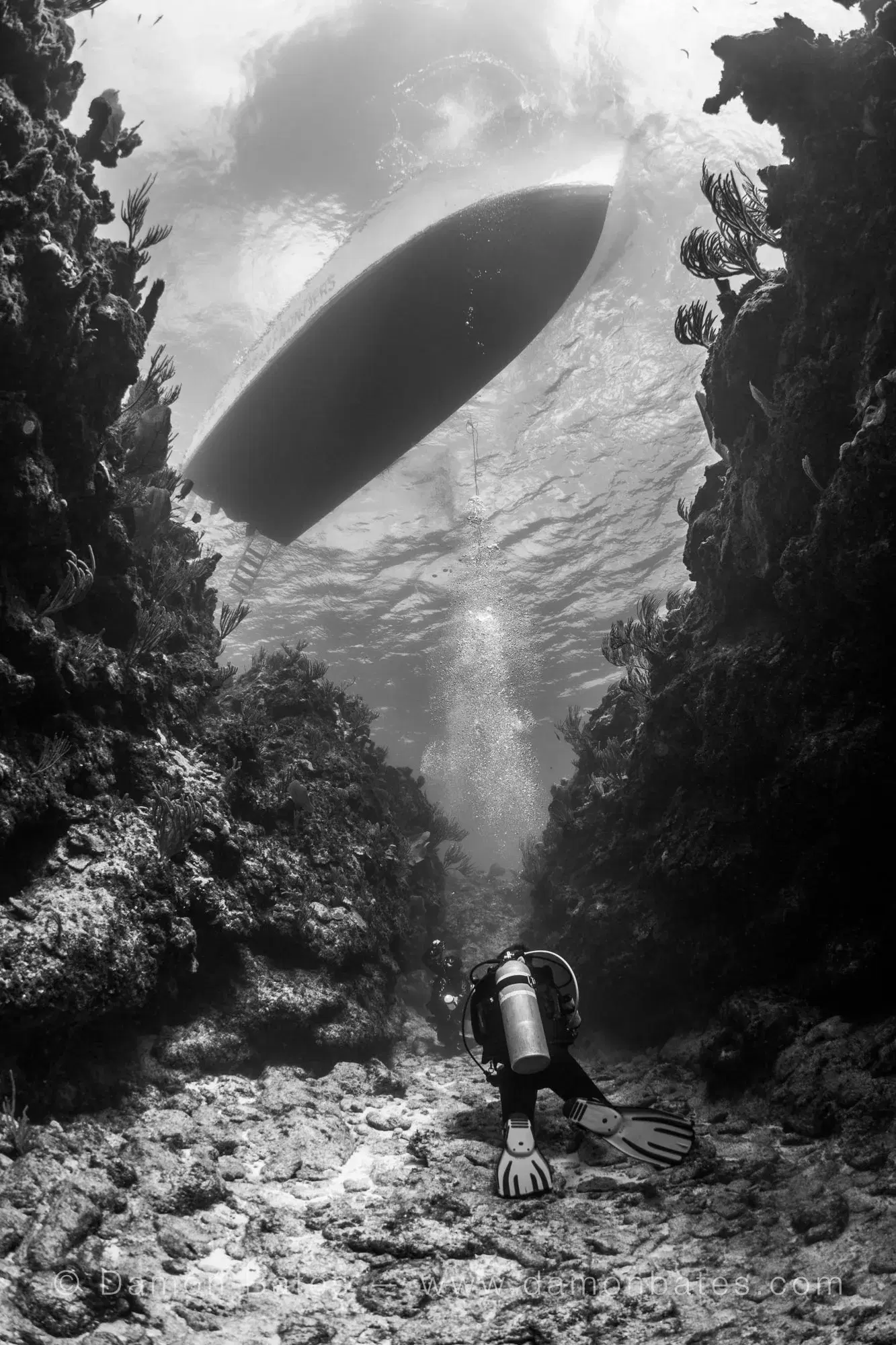 Scuba diver in a reef cut with a dive boat visible at the surface, Iron Shore Gardens, Grand Cayman — black and white — underwater photograph by Damon Bates