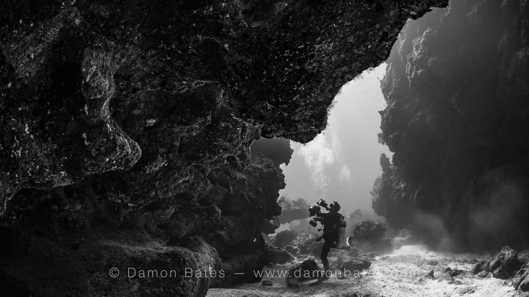 Underwater photographer with camera crouched in a cave arch at Iron Shore Gardens, Grand Cayman — black and white — underwater photograph by Damon Bates