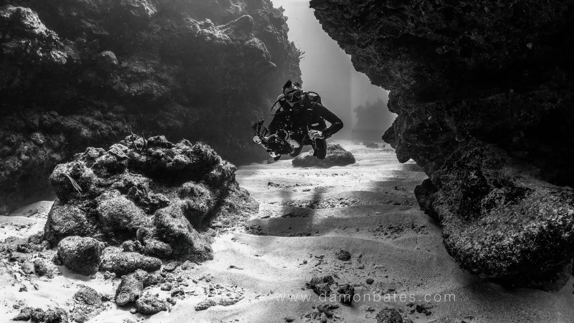 Scuba diver moving through a sandy corridor between rock walls at Iron Shore Gardens, Grand Cayman — black and white — underwater photograph by Damon Bates