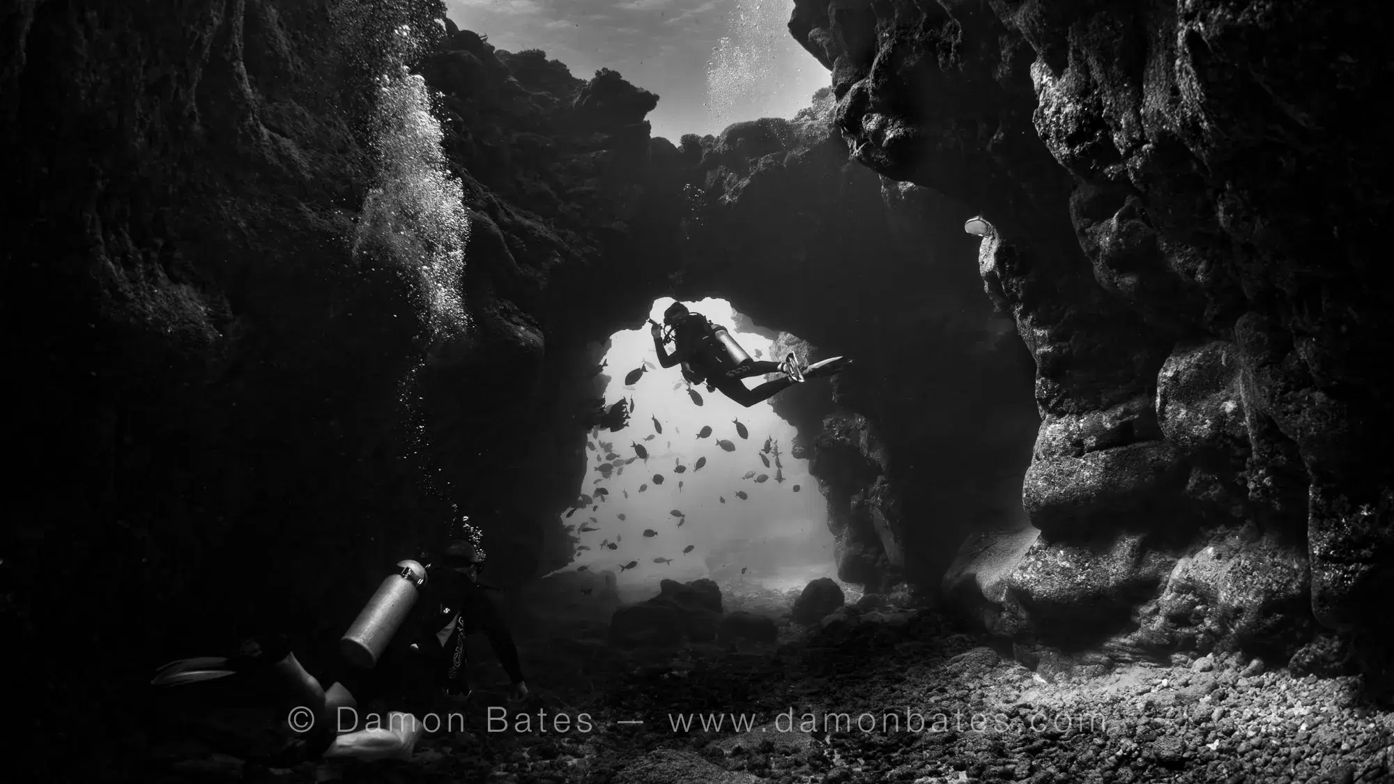 Two scuba divers framed in a lava arch at Sheraton Caverns, Kauai, Hawaii — black and white — underwater photograph by Damon Bates