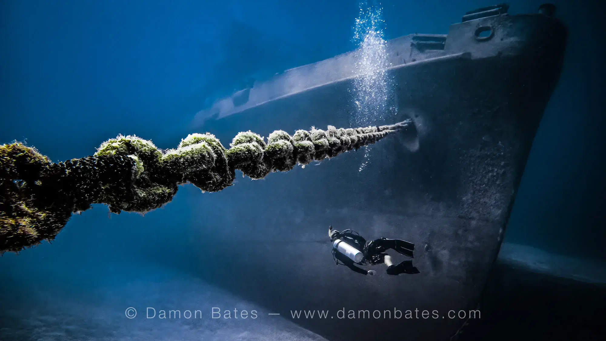 Scuba diver dwarfed by the coral-encrusted bow and anchor chain of the USS Kittiwake, Grand Cayman, 2016 — underwater photograph by Damon Bates