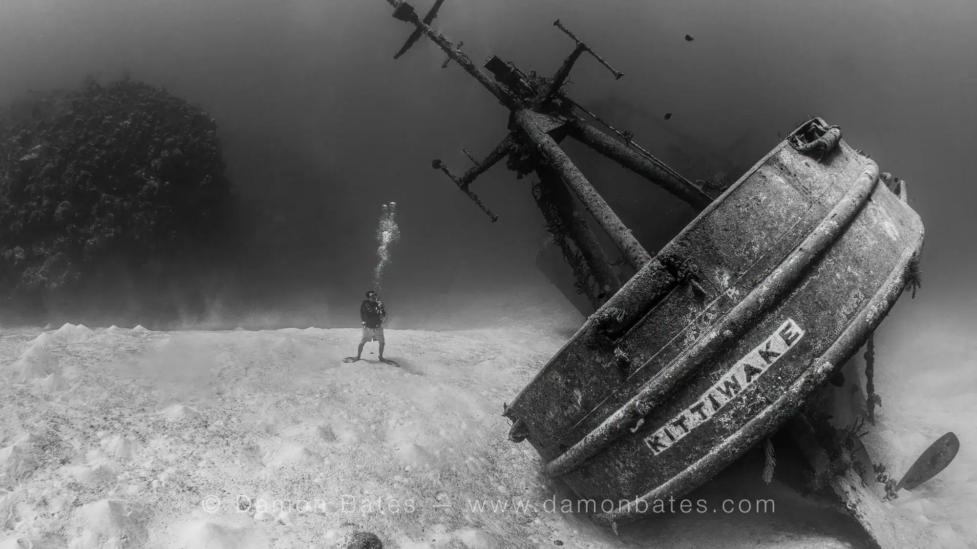 Scuba diver standing on the sandy seafloor beside the bow of the USS Kittiwake shipwreck, Seven Mile Beach, Grand Cayman, 2025 — underwater photograph by Damon Bates