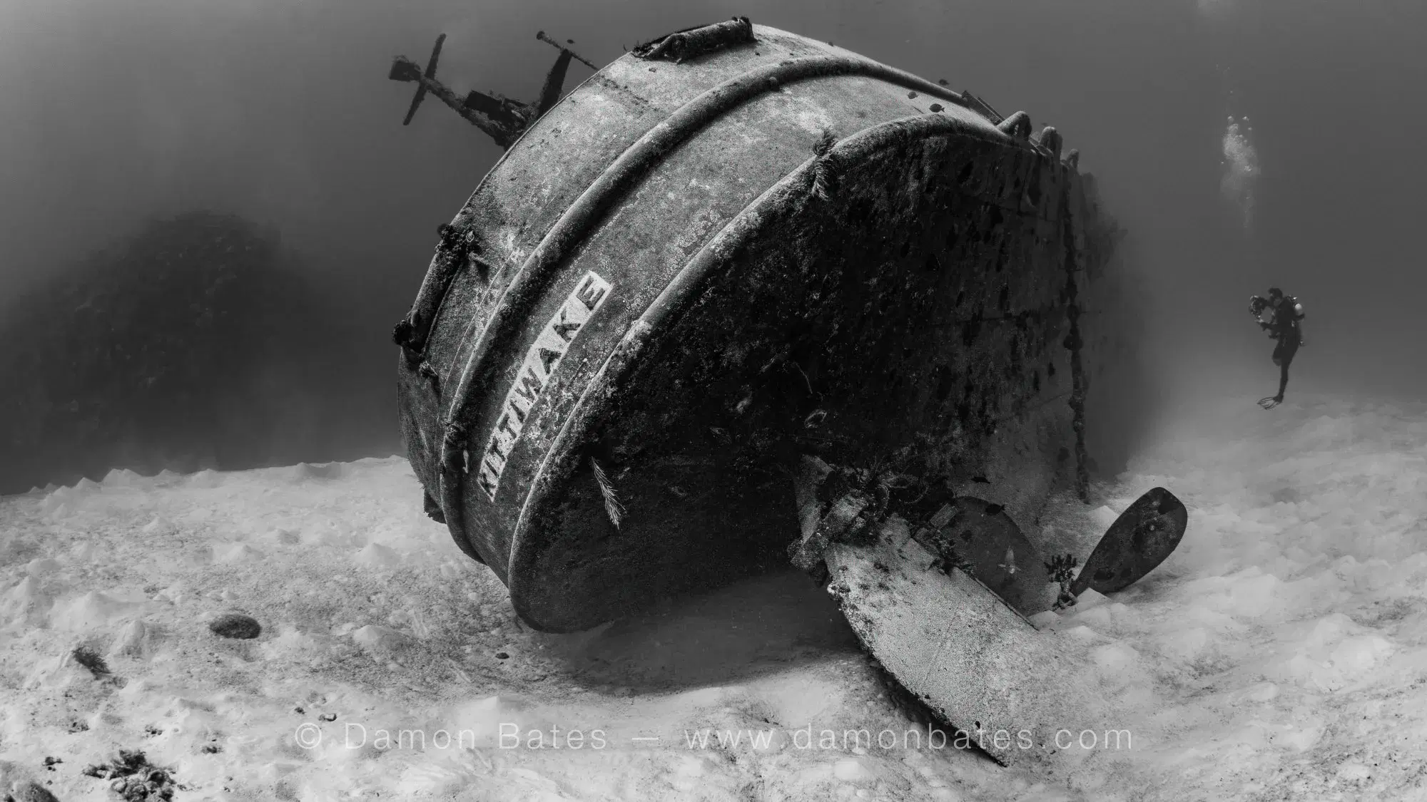 Stern of the USS Kittiwake with name visible on hull, diver in background, Seven Mile Beach, Grand Cayman, 2025 — black and white — underwater photograph by Damon Bates